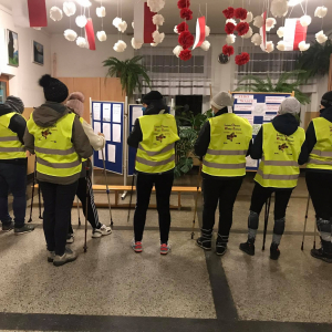 Uczestnicy zajęć z nordic walking. w holu  szkoły w tle dekoracja w biało czerwonych kwiatach z bibuły. Kliknięcie na zdjęcie spowoduje jego powiększenie do oryginalnego rozmiaru.
